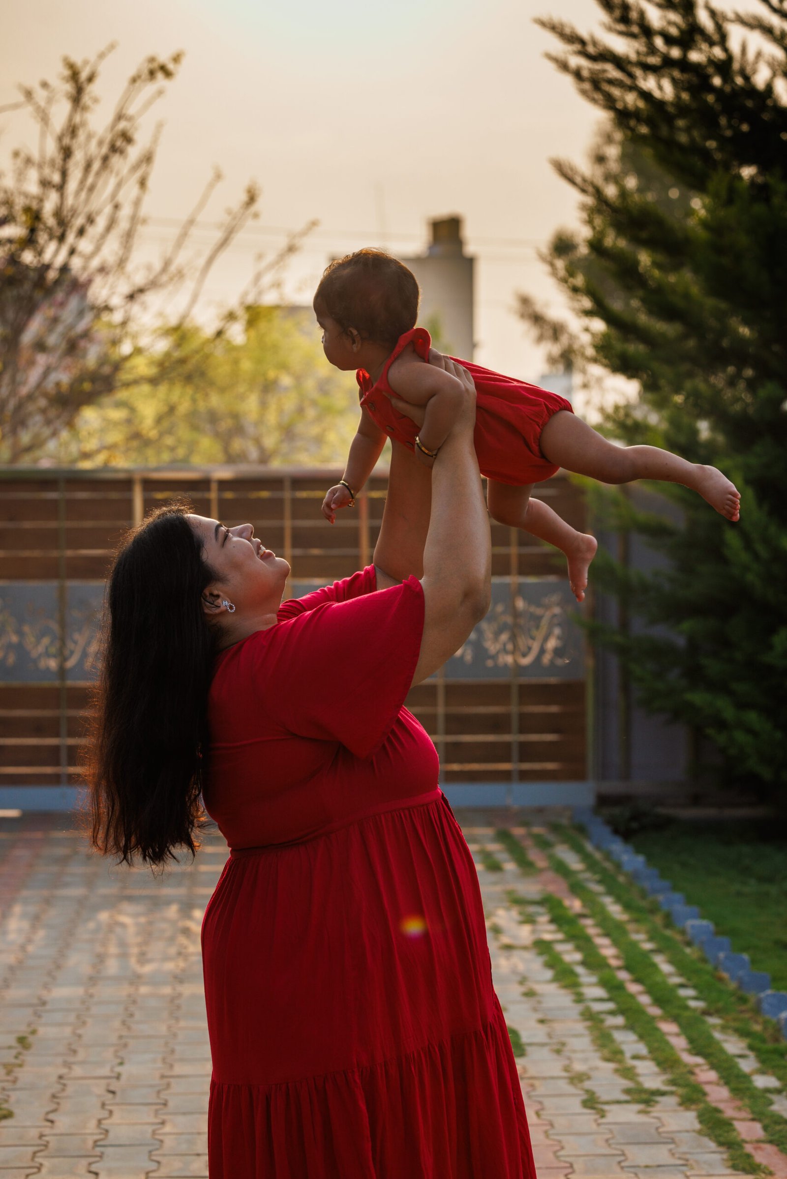 Mother in a red dress lifting her baby outdoors at sunset